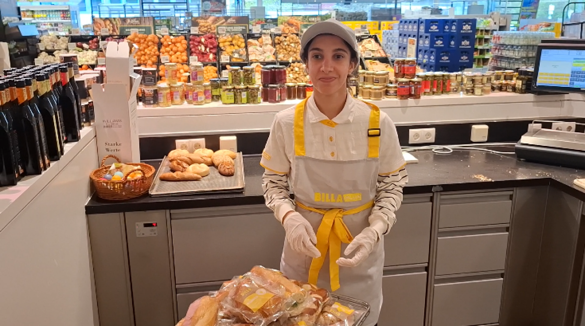 Junge Frau mit Arbeitskleidung von BILLA PLUS steht in einem Suoermarkt in der Feinkostzubereitung. Vor sich hat sie einen Rollwagen mit zubereiteten Weckerln stehen.