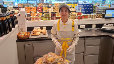 Junge Frau mit Arbeitskleidung von BILLA PLUS steht in einem Suoermarkt in der Feinkostzubereitung. Vor sich hat sie einen Rollwagen mit zubereiteten Weckerln stehen.