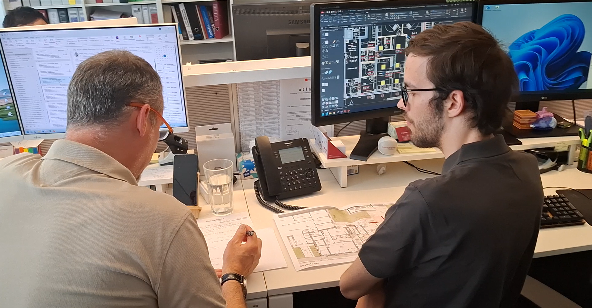 Zwei Männer von hinten fotografiert sitzen an einem Schreibtisch mit Computer-Bildschirmen. Der links sitzende erläutert dem rechts sitzenden etwas.