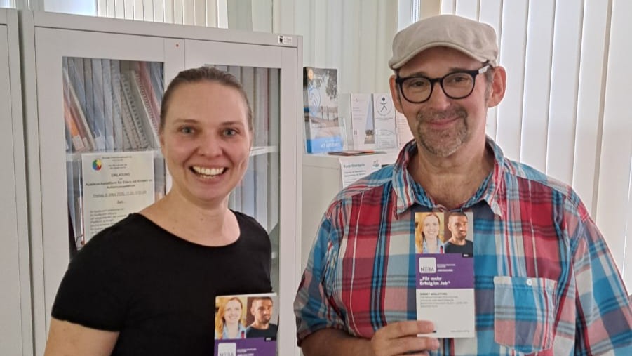Eine Frau in schwarzen T-Shirt und ein Mann in einem bunt karierten Hemd stehen in einem hellen Büro und halten lächelnd Faltfolder von NEBA in Händen.