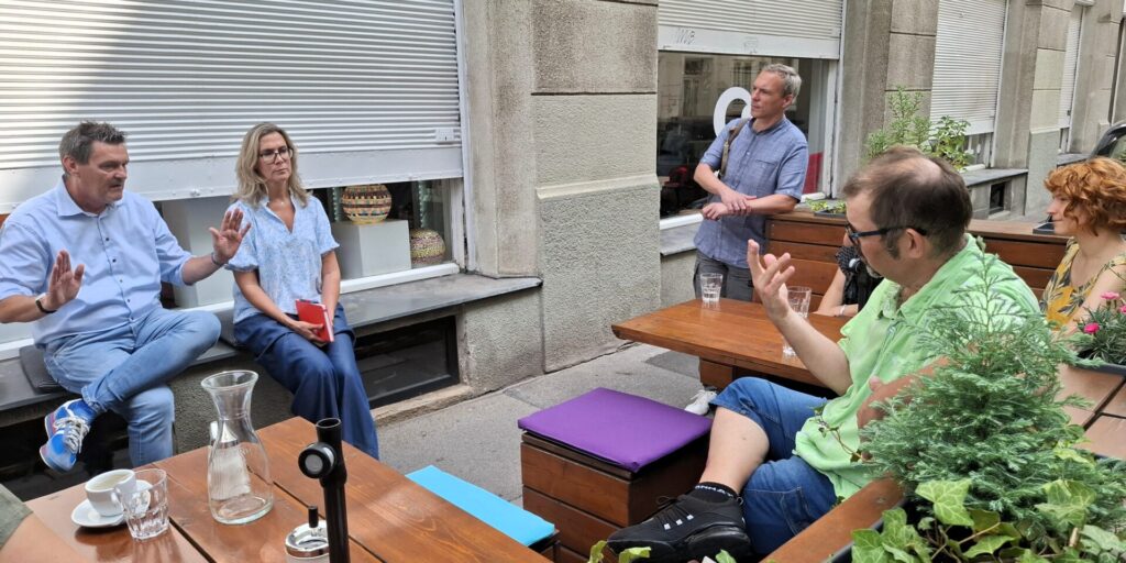 4 Personen sitzen in einem Straßengastgarten vor dem Geschäft der Kümmerei in Wien. Ein Mann und eine Frau sitzen am Fenstersims ihnen gegenüber. Zwei der Personen gestikulieren und sprechen im Moment des Fotos miteinander.