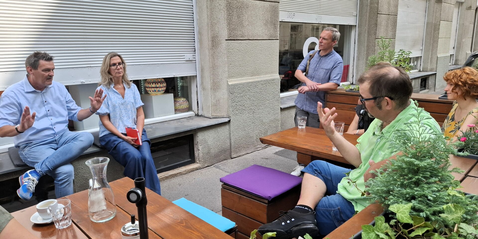 4 Personen sitzen in einem Straßengastgarten vor dem Geschäft der Kümmerei in Wien. Ein Mann und eine Frau sitzen am Fenstersims ihnen gegenüber. Zwei der Personen gestikulieren und sprechen im Moment des Fotos miteinander.