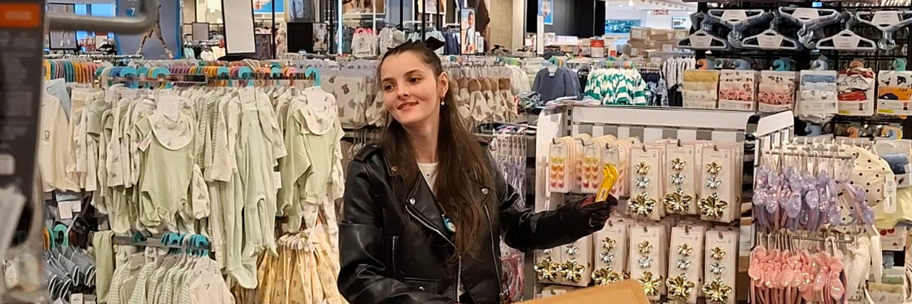 Junge Frau steht mit einem Pappkarton in der Hand auf der Verkaufsfläche eines Bekleidungsgeschäfts. In der linken Hand hält sie ein Cutter-Messer.