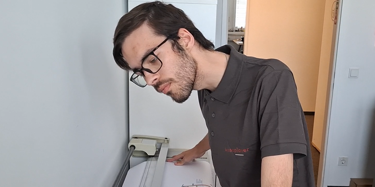 Mitarbeiter steht an einer großen Schnittmaschine für ausgedruckte Pläne und arbeitet konzentriert.
