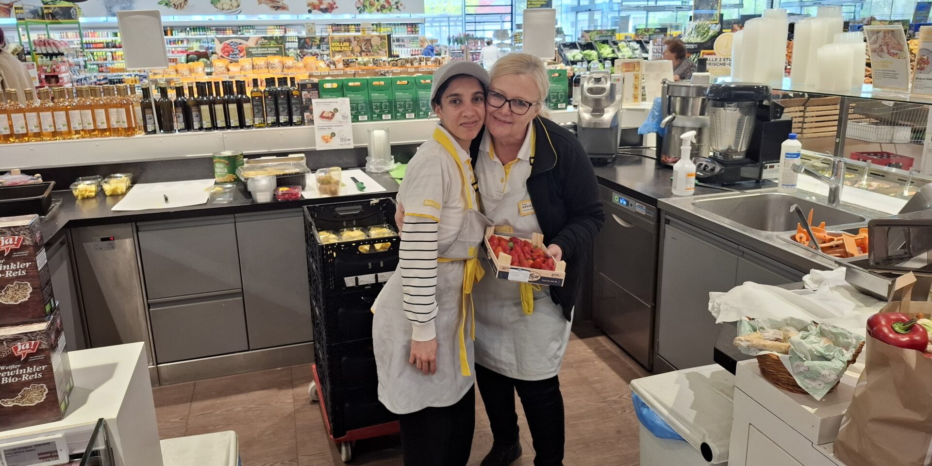 Mitarbeiterinnen der Feinkostabteilung eines Supermarkts stehen Arm in Arm nebeneinander.